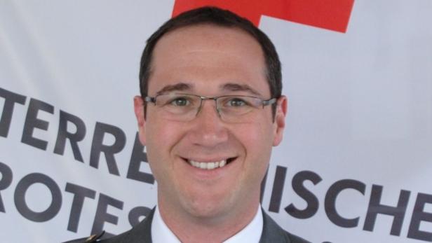 Ein lächelnder Mann mit Brille vor einem Banner des Roten Kreuzes.