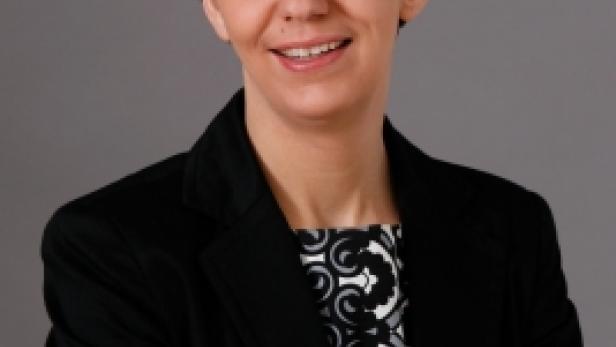 Eine Frau mit Brille und dunklem Blazer lächelt mit verschränkten Armen.