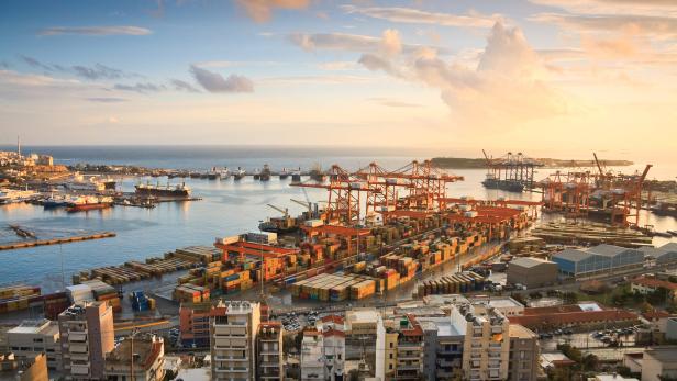 Blick auf einen Hafen mit zahlreichen Containern, Kränen und Schiffen bei Sonnenuntergang.