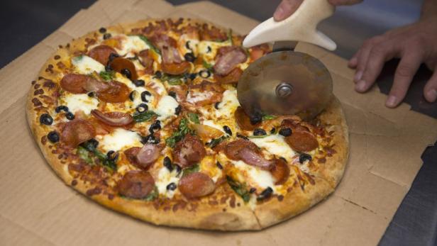 Eine Pizza mit Salami, Oliven, Zwiebeln und Spinat wird mit einem Pizzaschneider geschnitten.