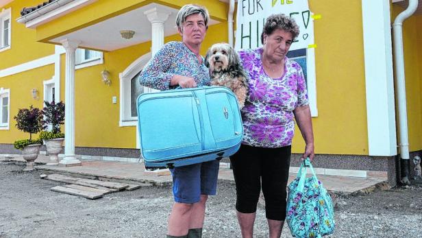 Zwei Frauen stehen mit einem Hund und Gepäck vor einem gelben Haus.