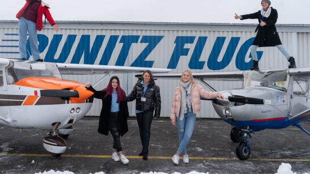 Fünf Frauen vor bzw. auf zwei Kleinflugzeugen vor einem Hangar