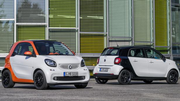 Zwei Smart-Autos, ein weiß-oranges Coupé und ein weiß-schwarzer Viertürer, stehen vor einem Gebäude.