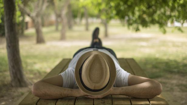 Restful boy wearing a straw hat laying down on a wooden table in the middle of the forrest at a park. You can put text on the background that is out of focus.