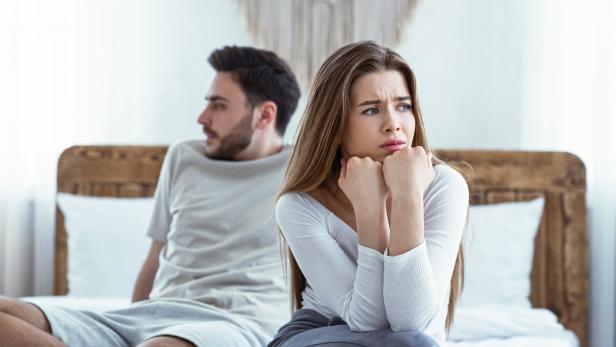 Mann und Frau sitzen getrennt auf einem Bett, die Frau wirkt besorgt.
