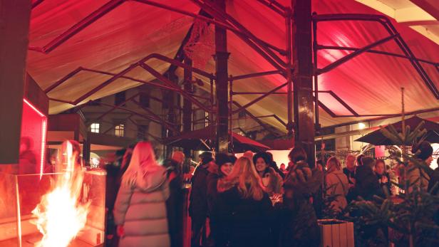 Menschen versammeln sich unter einem beleuchteten Zeltdach mit einer Feuerstelle im Vordergrund.