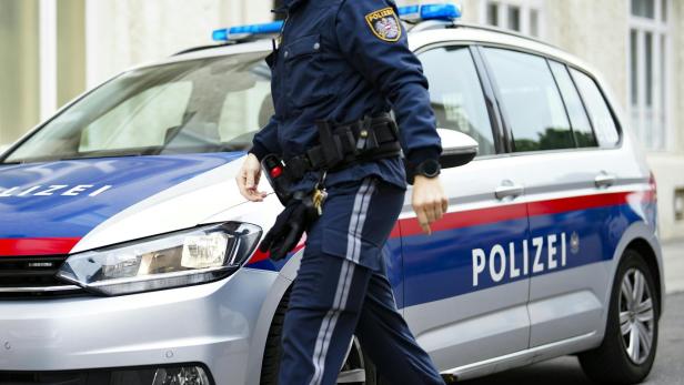 Ein Polizist in Uniform geht an einem silbernen Polizeiauto mit blauen und roten Streifen vorbei.