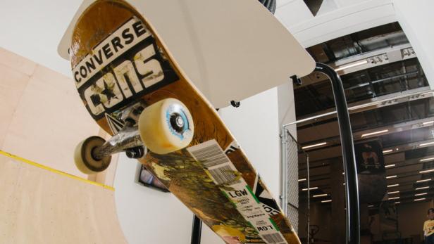 Ein Skateboard mit Converse-Logo steht auf einer Halterung, ein Skater im Hintergrund mit erhobenen Armen.