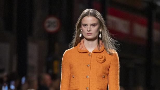 Ein Model präsentiert einen orangefarbenen Tweed-Mantel mit passendem Rock auf dem Laufsteg.