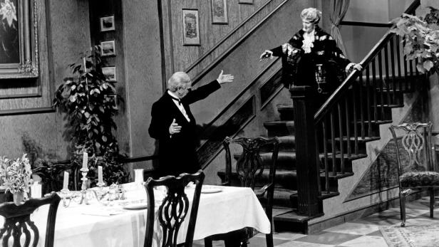 Ein Mann im Smoking begrüßt eine Frau auf einer Treppe in einem eleganten Haus.