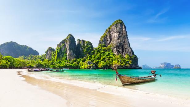 Ein traditionelles thailändisches Boot liegt an einem tropischen Strand mit Kalksteinfelsen im Hintergrund.