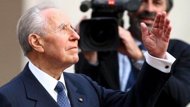 Ein älterer Mann in einem Anzug winkt in die Kamera.