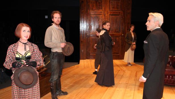 Szene aus einem Theaterstück mit mehreren Darstellern in historisch anmutenden Kostümen auf einer Holzbühne.
