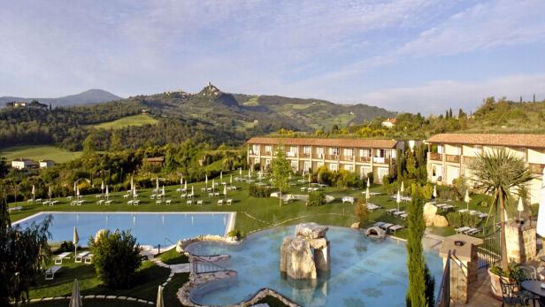 Ein Hotelresort mit Pools und Liegestühlen vor einer hügeligen Landschaft in der Toskana.