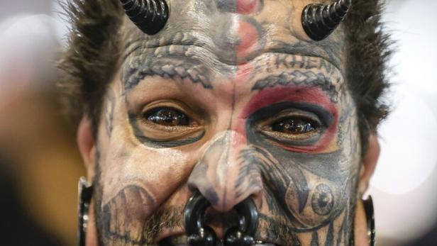 Ein Mann mit Gesichtstattoos, Hörnern auf der Stirn und Piercings in Nase und Lippen streckt die Zunge heraus.