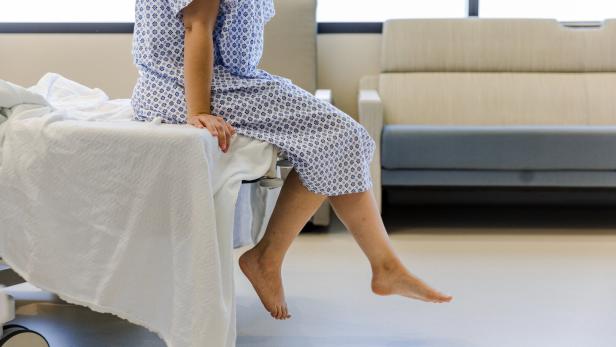 Frau in blau-weißem Operationsgewand sitzt barfuß auf einem Krankenbett 
