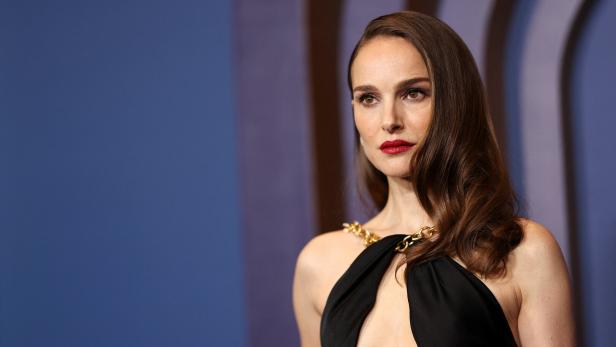 Natalie Portman in einem schwarzen Kleid mit goldenen Ketten vor einem blauen Hintergrund.