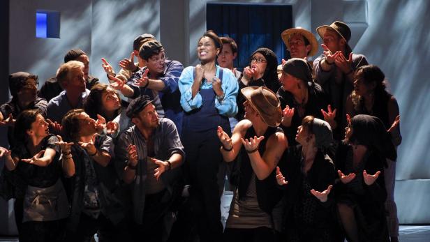 Eine Sängerin steht im Rampenlicht, umgeben von einem Chor auf einer Theaterbühne.