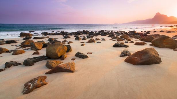 Ein sandiger Strand mit Felsen und einem Berg im Hintergrund bei Sonnenuntergang.