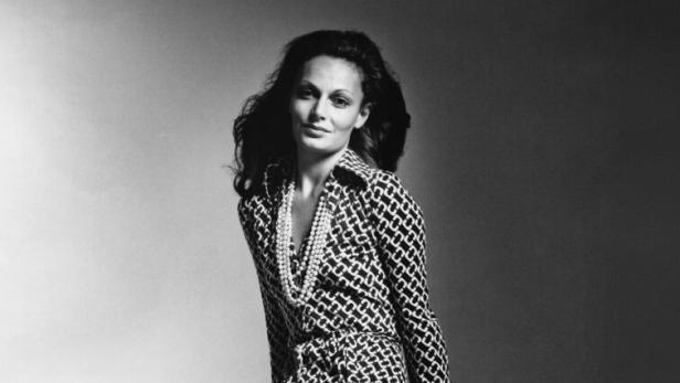 Eine Frau im gemusterten Kleid und mit Perlenkette posiert an einem weißen Würfel.