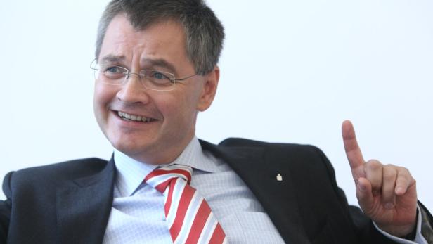 Ein lächelnder Mann mit Brille, Anzug und rot-gestreifter Krawatte hebt den Zeigefinger.