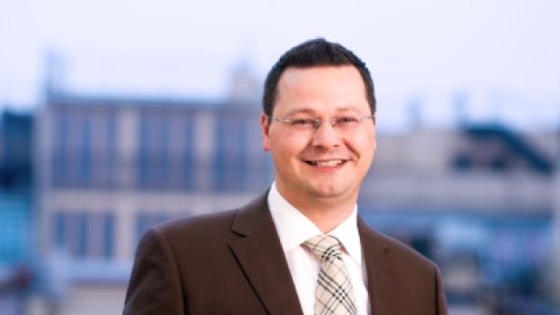 Ein lächelnder Mann mit Brille und Anzug vor einem Gebäude.