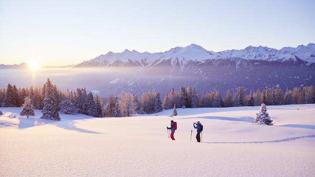 Zwei Personen wandern bei Sonnenaufgang durch eine verschneite Winterlandschaft mit Bergen im Hintergrund.