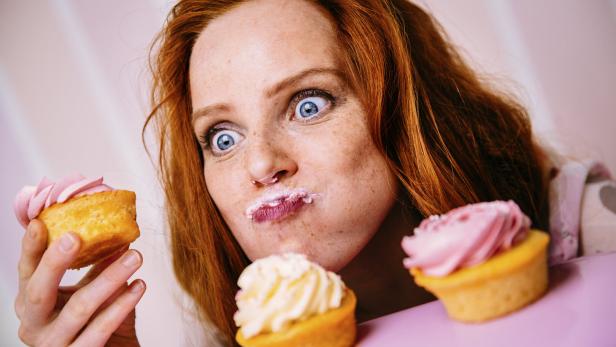 Eine rothaarige Frau mit Zuckerguss im Gesicht hält einen Cupcake und blickt mit großen Augen auf weitere Cupcakes.