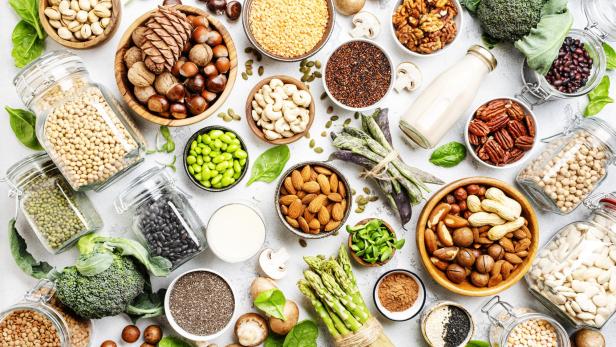 Verschiedene gesunde Lebensmittel wie Nüsse, Hülsenfrüchte, Gemüse und Samen sind auf einer Fläche angeordnet.