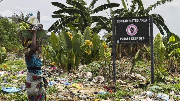 Ein Schild warnt vor Bodenverschmutzung durch Öl-Lecks, eine Frau trägt Bananen