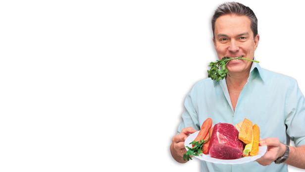 Ein lächelnder Mann hält einen Teller mit rohem Fleisch und Gemüse, Petersilie im Mund.