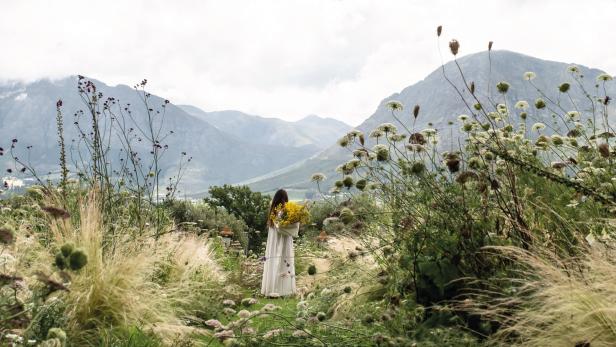 Eine Frau steht mit einem gelben Blumenstrauß in einer üppigen Landschaft vor einer Bergkulisse.