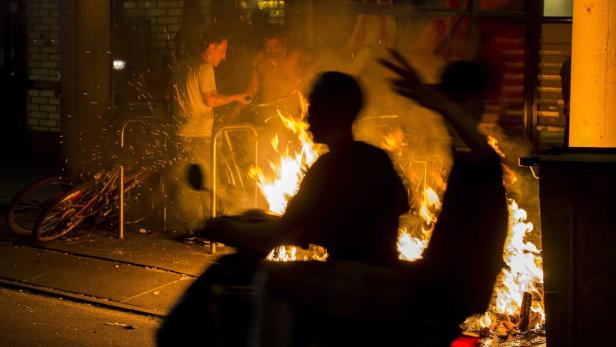 Mehrere Personen stehen nachts an einem Lagerfeuer in der Stadt.