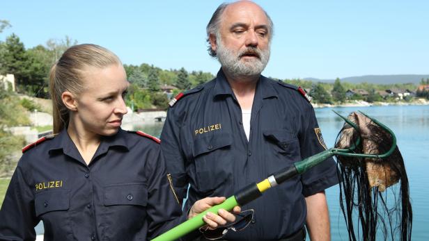 Zwei Polizisten halten einen Rechen mit einer nassen, schwarzen Perücke vor einem See.