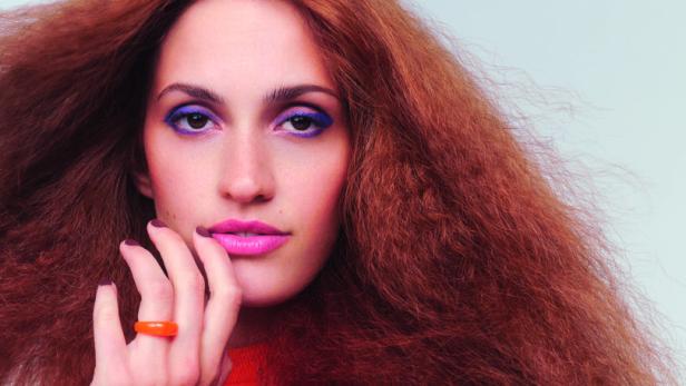 Eine Frau mit rotem, lockigem Haar und orangefarbenem Oberteil posiert vor einem alten Fernseher.