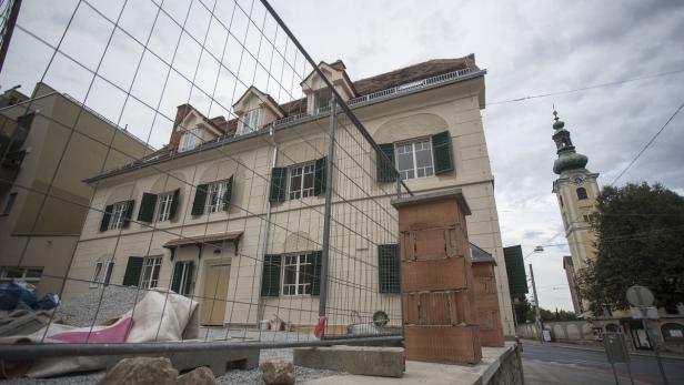 Ein renoviertes Gebäude mit grünen Fensterläden und einem Kirchturm im Hintergrund.