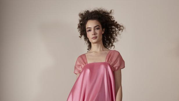 Eine Frau mit lockigem Haar trägt ein weites, rosafarbenes Kleid.