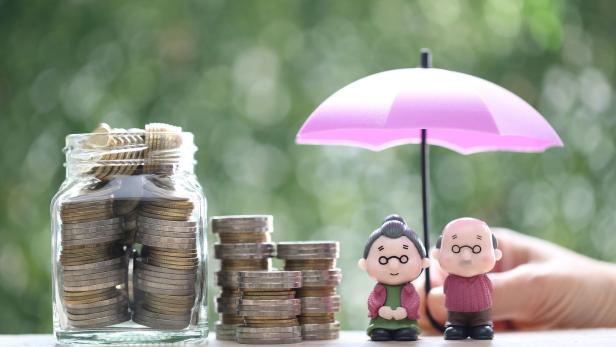 Zwei Spielzeugfiguren von einem älteren Pärchen stehen vor einer Hand, die einen Regenschirm hält. Daneben ein Stapel Münzen und Geld in einer Glasflasche auf grünem Hintergrund,