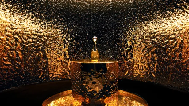 Ein goldener Parfümflakon steht auf einem goldenen Sockel vor einer goldenen Wand.