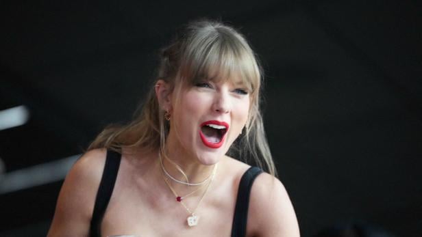 Taylor Swift mit offenem Mund und rotem Lippenstift, die ein schwarzes Top und mehrere Halsketten trägt.