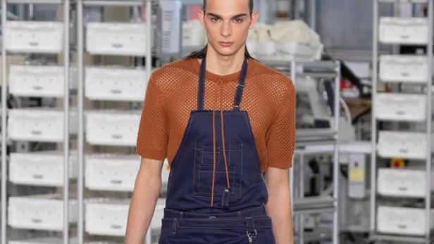 Ein Mann in einem orangefarbenen Netzhemd und einer blauen Latzschürze mit Shorts auf einem Laufsteg.