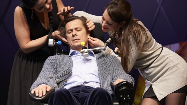 Zwei Frauen helfen einem Mann im Rollstuhl mit an seinem Kopf befestigten Geräten.