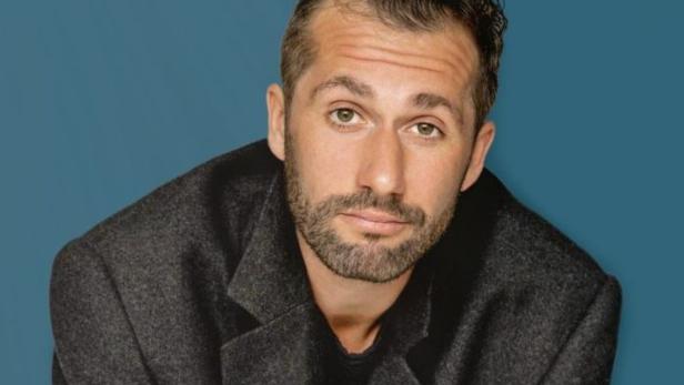 Ein Mann mit Bart und grauem Mantel vor einem blauen Hintergrund.