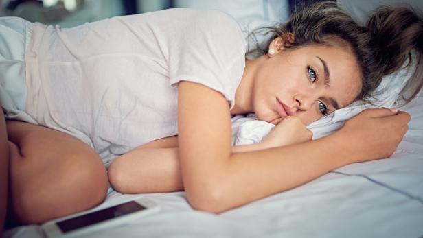 Junge Frau liegt nachdenklich auf einem Bett, den Kopf auf ein Kissen gestützt, ein Smartphone liegt neben ihr.