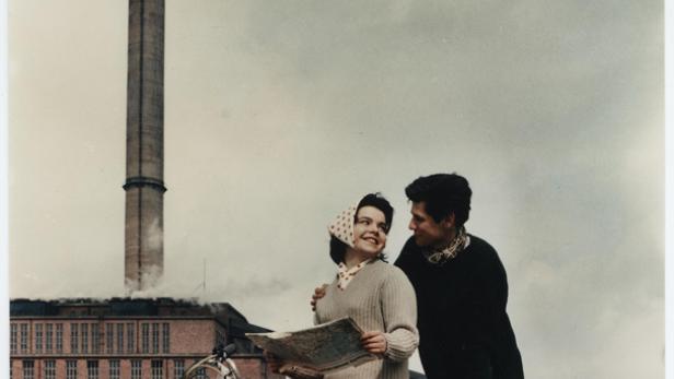 Ein junges Paar sitzt auf einem Motorroller mit einer Landkarte in der Hand vor einer Fabrik.