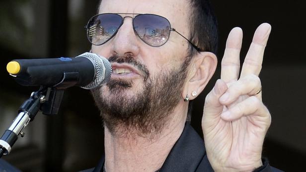 Ringo Starr mit Sonnenbrille am Mikrofon, der das Victory-Zeichen macht.