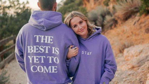 Ein Paar in lila Hoodies steht vor einer Felsformation; auf den Hoodies stehen die Worte „The best is yet to come“ und „Stay“.