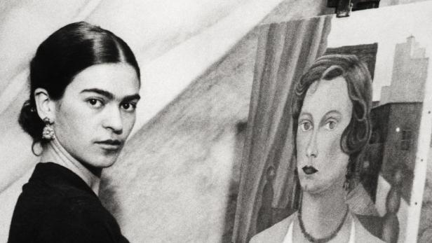 Frida Kahlo steht vor einer Staffelei und malt ein Porträt.