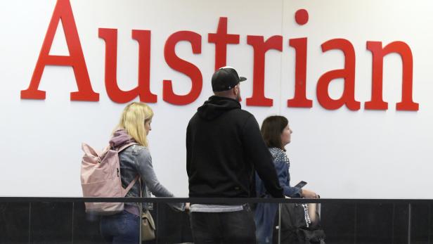 Reisende mit Gepäck vor einem roten „Austrian“-Schriftzug.