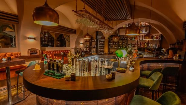 Eine Bar mit dunklen Holzmöbeln und grünen Samtstühlen unter warmem Licht.